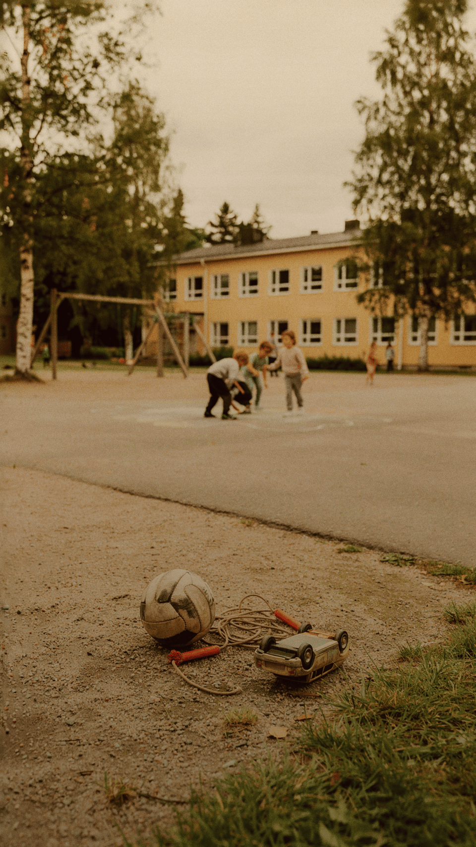 Leksaker på en skolgård i förgrunden med barn som leker i bakgrunden vid en gul skolbyggnad.