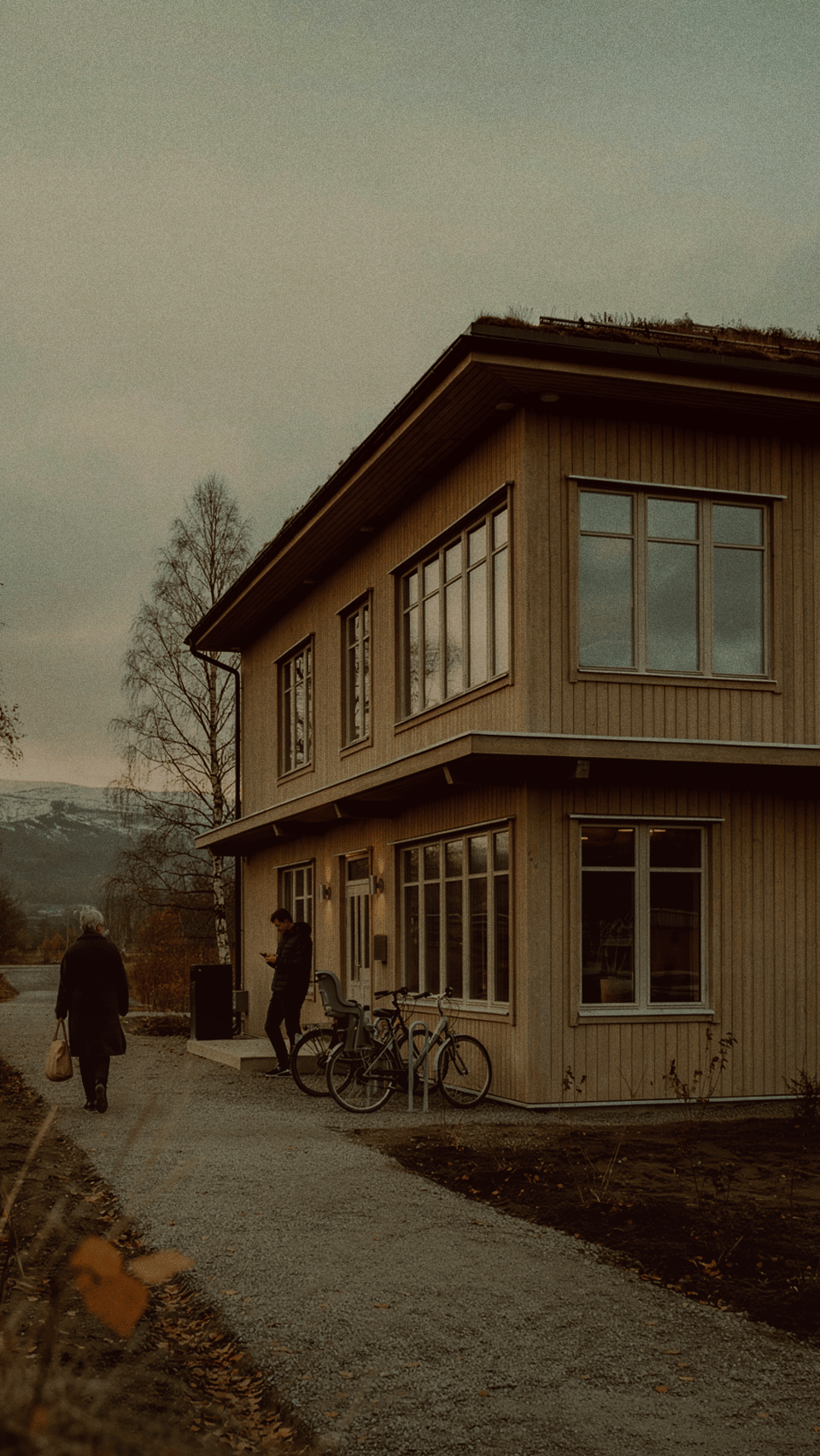 En modern träbyggnad med sedumtak i en glesbygdsmiljö. En person går på en grusväg medan en annan står vid parkerade cyklar utanför entrén.