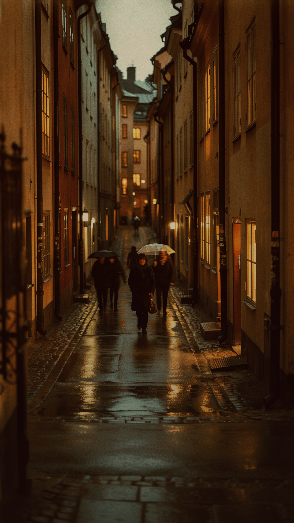 Människor med paraplyer promenerar längs en smal, våt kullerstensgata i Stockholm under kvällstid med varmt ljus från fönster.